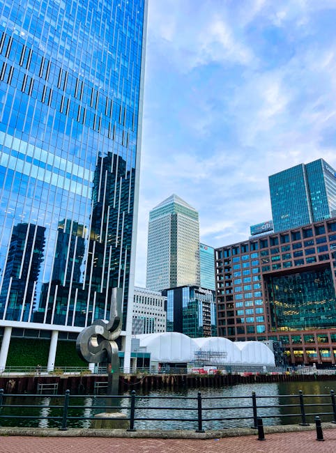 A black-and-white photograph of a modern urban skyline showing a range of high-rise buildings along a waterfront. The image features tall skyscrapers with reflective glass facades, some with distincti