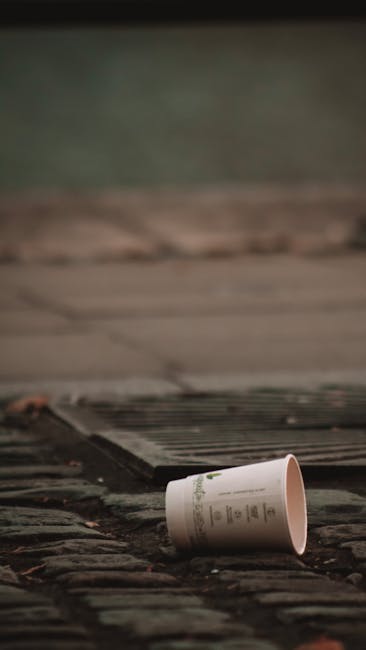 A man with dark hair, dressed in a black T-shirt with white and blue text, is seen leaning over a metallic public rubbish bin on a paved outdoor walkway. He is holding a large white plastic bag filled